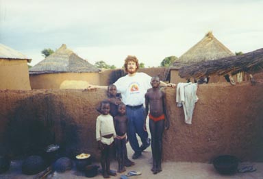 Bawku, Ghana, 1994. Peace Corps volunteer Wayne Breslyn stands with several Kusasi children in their village. On the left of the picture is the area where meals are prepared. Photo by Peace Corps Volunteer Wayne Breslyn.